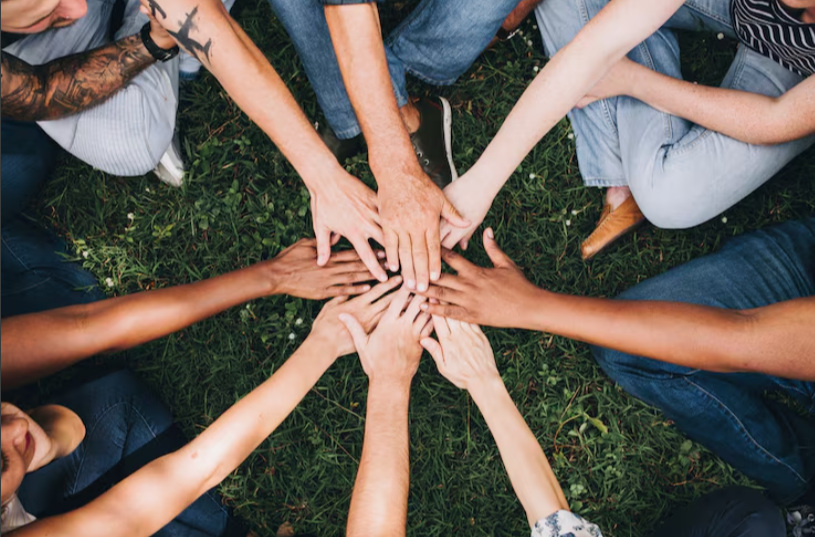 Several people sitting in a circle on grass, reaching their hands into the centre and placing them together.